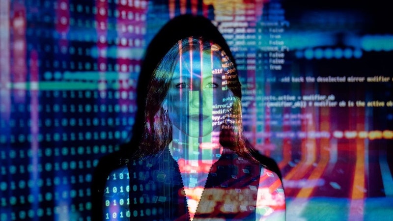 Fotoğrafın ortasında yüzü bulanık görünen bir kadın görülüyor. Beyaz bir tişört giymiş ve saçlarını arkaya toplamış. Yüzü hafifçe odak dışında, ancak gözleri hala tanınabilir durumda. Resmin sol tarafında bulanık bir kod görüntüsü var. Sağ tarafta, bir dizi simgenin göründüğü bir bilgisayar ekranının yakın çekimi var. Daha sağda, alt köşede sayıların yakın çekimi var. Arka plan bulanık bir bayrak görüntüsüyle doldurulmuş. Fotoğrafın sıcak, sarı bir tonu var.