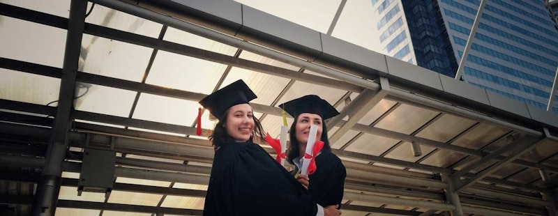 Her ikisi de siyah mezuniyet cübbesi ve kepi giymiş iki kadın yan yana duruyor. İlk kadın elinde iki diploma tutuyor ve kepi açık gri renkteyken, ikinci kadının kepi daha koyu bir siyah. Her iki kadın da mutlu bir şekilde gülümsüyor, gururlu ve başarılı görünüyor. İlk kadının cübbesi düz siyah renktedir, ikinci kadının cübbesinde ise altın süslemeli beyaz bir kuşak vardır. İkinci kadının koyu renk şapkasının kenarından sarkan altın bir püskül vardır. Her iki kadın da kulaktan kulağa sırıtıyor ve dünyayı ele geçirmeye hazır görünüyor.