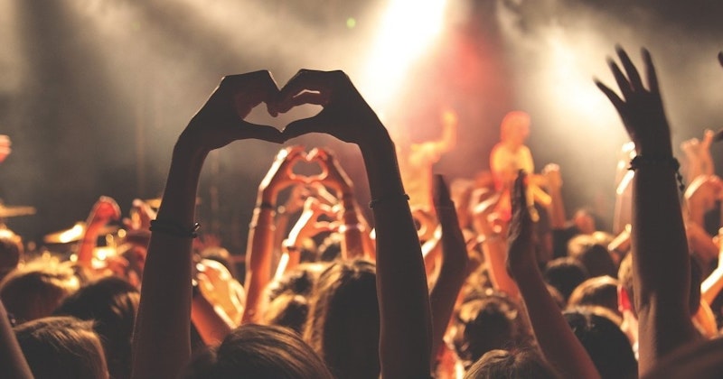 Büyük bir grup insan kalp şeklinde bir araya toplanmış, ellerini başlarının üzerinde birleştirmiş. Grubun çoğunluğu net bir şekilde odaklanmışken, kalabalığın kenarları bulanık ve odak dışıdır. Kalp şeklinin ortasındaki kişinin bileğinde bir bilezik var. Daha solda, boynundan aşağı saç telleri dökülen birinin saçına yakın çekim yapılmış. Sağda bir başka kişinin kafasının bulanıklığı görülüyor ve daha sağda başka bir kişinin yüzünün yakın çekimi var, gözleri konsantrasyon içinde kapalı. Görüntü renkli ve canlı; grubun parlak kıyafetleri ve gülümsemeleri neşeli bir atmosfer yaratıyor.