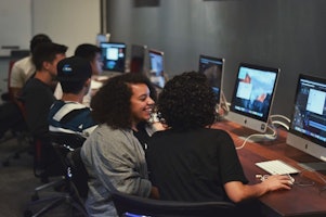 Bir grup insan birden fazla bilgisayarın bulunduğu bir masanın etrafında oturmaktadır. Çizgili gömlek giyen bir kişinin önünde açık bir dizüstü bilgisayar var. Başka bir kişi başını eğmiş, yüzü görünmüyor ama kıvırcık saçları var ve gülümsüyor. Üçüncü bir kişi ise üzerinde mavi beyaz bir logo bulunan siyah bir şapka takıyor. Masa bilgisayar klavyeleriyle doludur ve birkaç ekran bulanık görünmektedir. Masadaki herkes önlerindeki bilgisayarlara odaklanmış gibi görünüyor.