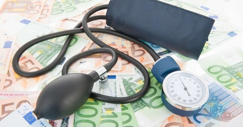 Bir yığın paranın üzerinde duran bir tonometre ve siyah bir stetoskopun yakın çekimi. Tonometre kan basıncını ölçmek için kullanılan tıbbi bir cihaz, siyah stetoskop ise tıp uzmanları tarafından kalp ve akciğerleri dinlemek için kullanılan bir alet. Resmin sol tarafında, içinden birkaç banknot para çıkan siyah bir cüzdan görülüyor. Sağ alt köşede, duvarda küçük, bulanık bir saat asılı. Görüntü hafif sarımsı bir renk tonuna sahip ve tonometre ile para metalik bir parlaklığa sahip.