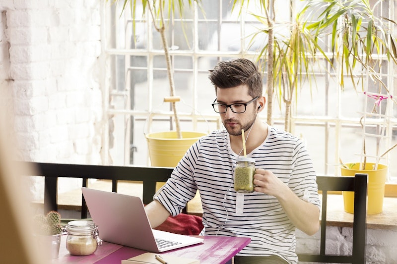 Gözlüklü ve beyaz gömlekli bir adam, önünde dizüstü bilgisayarıyla bir masada oturuyor. Kapaklı bir cam kavanozdan yeşil bir smoothie içmektedir. Yüzü yakın plandadır, gözlükleri ve saçının birkaç teli görünmektedir. Yan tarafında, siyah saplı sarı bir kova var. Dizüstü bilgisayarına odaklanmış, görünüşe göre bir iş üzerinde çalışıyor. Smoothie'si ve sarı kova, normalde nötr olan sahneye biraz renk katıyor.
