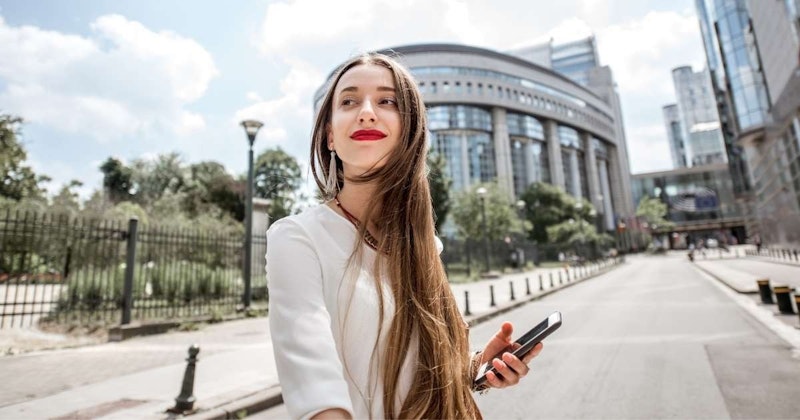 Kırmızı ruj sürmüş, uzun kahverengi saçlı bir kadın uzun beyaz bir binanın önünde durmakta ve sağ elinde siyah bir telefon tutmaktadır. Üzerinde açık sarı desenli açık mavi bir gömlek var ve yüzünde nazik bir gülümseme var. Uzun saçları rüzgârda savruluyor ve omuzlarına kadar uzanıyor. Görüntünün arka planı hafif bulanık ve köşede siyah bir nesne var. Kadın, ön planda siyah bir çit bulunan uzun beyaz bir binanın önünde duruyor. Görüntü bir sıcaklık ve neşe hissiyle doludur.