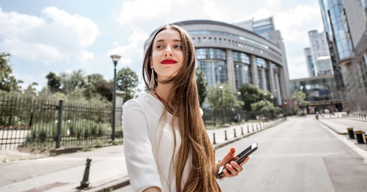 Kırmızı ruj sürmüş, uzun kahverengi saçlı bir kadın uzun beyaz bir binanın önünde durmakta ve sağ elinde siyah bir telefon tutmaktadır. Üzerinde açık sarı desenli açık mavi bir gömlek var ve yüzünde nazik bir gülümseme var. Uzun saçları rüzgârda savruluyor ve omuzlarına kadar uzanıyor. Görüntünün arka planı hafif bulanık ve köşede siyah bir nesne var. Kadın, ön planda siyah bir çit bulunan uzun beyaz bir binanın önünde duruyor. Görüntü bir sıcaklık ve neşe hissiyle doludur.