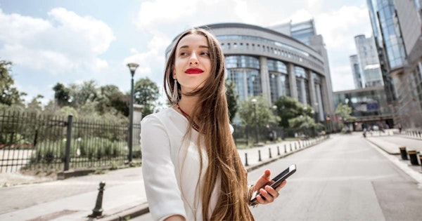 Kırmızı ruj sürmüş, uzun kahverengi saçlı bir kadın uzun beyaz bir binanın önünde durmakta ve sağ elinde siyah bir telefon tutmaktadır. Üzerinde açık sarı desenli açık mavi bir gömlek var ve yüzünde nazik bir gülümseme var. Uzun saçları rüzgârda savruluyor ve omuzlarına kadar uzanıyor. Görüntünün arka planı hafif bulanık ve köşede siyah bir nesne var. Kadın, ön planda siyah bir çit bulunan uzun beyaz bir binanın önünde duruyor. Görüntü bir sıcaklık ve neşe hissiyle doludur.