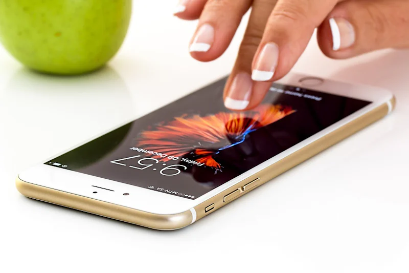 Bir kişi yakın planda, parmağıyla bir cep telefonunun ekranına dokunurken görülüyor. Telefon kişinin elinde ve yüzüne yakın bir konumda tutulmaktadır. Ekran yeşil bir elma simgesiyle aydınlatılmıştır ve kişi parmak ucuyla ekrana bastırmaktadır. Kişi cihazla etkileşime girerken odaklanmış bir ifadeye sahiptir. Görüntü net ve berraktır, telefonun ve kişinin parmağının ayrıntılarını gösterir.