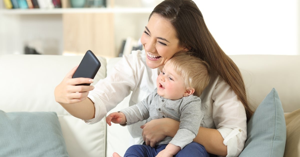 Genç bir kadın kucağında küçük bir bebekle selfie çekiyor. Kadın kameraya gülümsüyor, yüzü sevinçle aydınlanıyor. Kolu kadının boynuna dolanmış olan bebek de gülümsüyor. Hem anne hem de çocuk pembe desenli beyaz üstler giyiyor. Arka planda, şekli ve dokusu açıkça belli olan bir yastık görülüyor. Bir elin siyah dikdörtgen bir nesneyi tuttuğu görülürken, sağ alt köşede siyah bir nesnenin yakın çekimi görülüyor. Son olarak, bir el, yüzeyi ışığı yansıtan bir telefonu tutarken görülüyor. Genel olarak bu, bir anne ile çocuğu arasındaki mutlu ve samimi bir an.