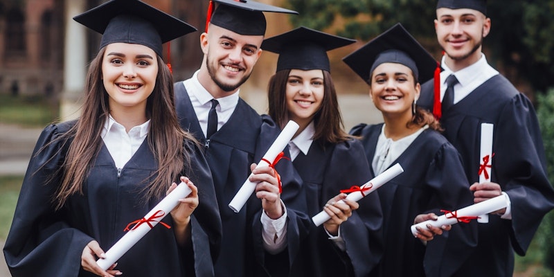 Mezuniyet cübbesi ve kepi giyen bir grup insan bir odada bir arada durmaktadır. Bir kadın önde siyah bir cüppe giymiş, beyaz bir kep takmış ve elinde beyaz bir diploma tutmaktadır. Onun arkasında bir grup insan da mezuniyet cüppesi ve kepi giymiş ve hepsinin elinde diploma var. Daha geride bir kişi mezuniyet kepi takmış ve elinde bir diploma tutuyor. Resmin sağ tarafında bir kişi diploma tutuyor ve daha geride başka bir kişi diploma tutuyor. Ön planda bir vazo içinde kırmızı bir çiçek var. Başarılarını kutlarken herkesin yüzünde ciddi ve gururlu bir ifade var.