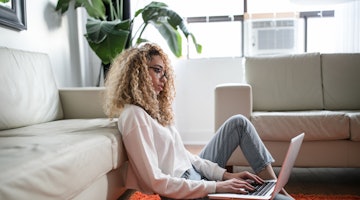 Kıvırcık sarı saçlı ve gözlüklü bir kadın kucağında dizüstü bilgisayarıyla yerde oturmaktadır. Elini dizüstü bilgisayarın üzerine koymuş ve ekrana bakıyor. Üzerinde beyaz bir tişört ve siyah bir kot pantolon var. Etrafındaki alan çoğunlukla beyaz, beyaz bir yastık ve etrafa saçılmış birkaç başka eşya var. Görüntü yakın çekimdir ve kadın ile dizüstü bilgisayarının samimi bir görüntüsünü verir. Bu görüntü, kadının faaliyetini ve çevresini net bir şekilde yansıttığı için bir görüntü başlığı veri kümesi hazırlamak için mükemmeldir.