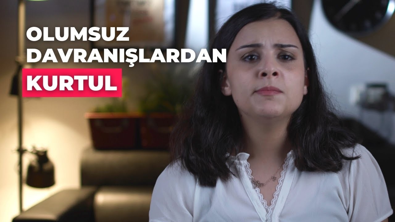 Uzun siyah saçlı, beyaz gömlekli bir kadın bulanıklaştırılmış bir kanepenin önünde durmaktadır. Ellerini kalçalarına koymuş ve kameradan uzağa bakmaktadır. Kanepe çizgili bir desene sahip ve çeşitli dekoratif yastıklarla dolu. Kanepenin sağ tarafında küçük, bulanık bir kedi uyurken görülüyor. Kadının yüzünde ciddi ve kararlı bir ifade var. Görüntünün arka planı nötrdür ve başka hiçbir nesne görünmemektedir.