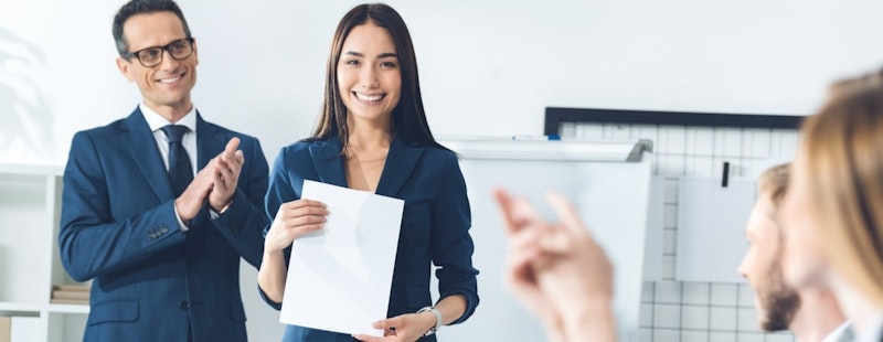 Bir kadın beyaz bir arka planın önünde ayakta durmakta ve elinde bir kağıt parçası tutmaktadır. Mavi bir bluz ve siyah bir etek giymiştir. Saçları düzgün bir topuzla bağlanmış ve kâğıda bakıyor. Yüzünde ciddi bir ifade var. Kolları hafifçe bükülmüş ve elleri kâğıdı sıkıca kavramış. Kâğıt beyaz ve üzerinde siyah bir yazı var. Sağ tarafında gri takım elbiseli ve mavi kravatlı bir adam var. Adam coşkulu bir ifadeyle ellerini çırpıyor. İkisinin de arkasında üzerinde yazılar olan bir beyaz tahta var. Kadın kâğıda, adam da beyaz tahtaya odaklanmış, ikisi de işleriyle meşgul.