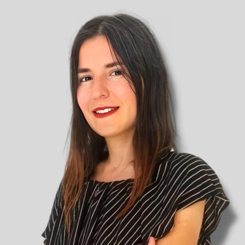 Uzun dalgalı siyah saçlı genç bir kadın beyaz bir arka plan üzerinde duruyor. Üzerinde ince siyah beyaz çizgili bir gömlek ve mavi bir kot pantolon var. Ellerini kalçalarına koymuş, kendinden emin ve kararlı bir ifadeyle doğrudan kameraya bakıyor. Gözlerinin kenarları kırışan sıcak bir gülümsemesi var. Saçları geriye doğru toplanmış ve birkaç tutam yüzünü çerçeveliyor. Duruşu rahat ama kendinden emin, güçlü ve bağımsız bir varlık sergiliyor.