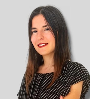 A young woman with long wavy dark hair stands against a white background. She is wearing a striped shirt with thin black and white stripes, and a pair of blue jeans. Her hands are placed on her hips, and she is looking directly into the camera with a confident and determined expression. She has a warm smile with her eyes crinkling in the corners. Her hair is pulled back with a few strands framing her face. Her posture is relaxed yet confident, displaying a strong and independent presence.