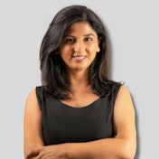 This is an image of a woman with dark black hair tied up in a bun. She has her arms crossed over her chest and is looking away from the camera. She wears a black top with long sleeves and a white bottom. Her facial expression is serious and her eyes are hidden from view. She stands in front of a white wall with nothing else in the image. She appears to be deep in thought, and her body language suggests she is feeling contemplative.