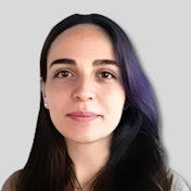 This photo features a woman with dark, shoulder-length hair. She is looking away from the camera and has a slightly serious expression on her face. She is wearing a plain white shirt and has her hands folded in front of her. The background is a light grey colour and there are no other objects in the frame. She is standing in a neutral pose with her feet slightly apart and her arms hanging down. The overall atmosphere of the photo is calm and peaceful.