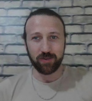 A middle-aged man with a thick, dark beard is standing in front of a cream-colored wall. He has a serious expression on his face and is wearing a black shirt. His beard is neatly trimmed and extends down towards his chest. His eyes are looking directly at the camera, and he has a slight smile on his face. His head is slightly tilted to the side and his hair is combed neatly. He is standing with his hands resting at his sides.