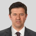 Yakışıklı, iyi giyimli bir adam profesyonel bir pozda duruyor. İki parçalı siyah bir takım elbise ve bembeyaz bir gömlek giymiş, desenli koyu renk bir kravatla tamamlamış. Siyah ayakkabıları mükemmel bir şekilde parlatılmış ve saçları düzgünce şekillendirilmiş. Duruşu kendinden emin ve doğrudan kameraya bakıyor. Yirmili yaşlarının sonunda ya da otuzlu yaşlarının başında görünüyor.