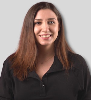 A young woman is smiling brightly and looking directly towards the camera. She has long, dark brown hair, which is partially tucked behind her ears. Her eyes are full of life and her lips are curved into a soft, genuine smile. She is wearing a white t-shirt, light blue jeans, and black shoes. Her expression is cheerful and her posture is relaxed. She is radiating confidence and joy, making her a beautiful and uplifting sight. She is standing in a bright, sunny environment and her warm glow is accentuated by the sun's rays.