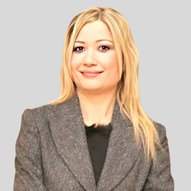 Dalgalı, sarı saçlı güzel bir kadın şık, gri bir takım elbise giyiyor. Yüzünde kendinden emin bir ifade var, gözleri doğrudan kameraya bakıyor. Kollarını göğsünün üzerinde kavuşturmuş ve uzun bacaklarını ayak bileklerinden kavuşturmuş. Bir çift siyah, sivri burunlu topuklu ayakkabı giyiyor. Boynunda inci bir kolye ve bir çift küçük altın küpe var. Dudakları açık pembe tonuna boyanmış. Dengeli ve zarif görünüyor.