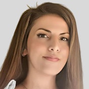 This is a close-up shot of a young adult woman with tan skin and shoulder-length wavy brown hair. She is looking off to the left with a slight smile, her eyes lightly closed. Her brows are slightly furrowed, giving her a thoughtful expression. Her chin and jawline are strong, and she has a few freckles scattered across her cheeks. She is wearing a light pink shirt with a white collar.