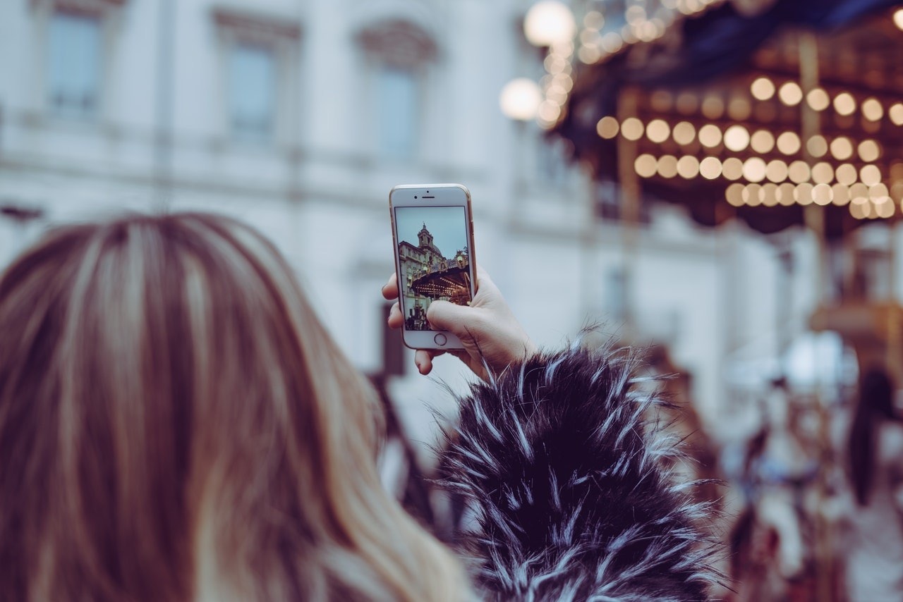 Bir kadın bir binanın önünde durmuş, telefonuyla selfie çekiyor. Telefonu sağ eliyle tutuyor ve sol eli havada. Beyaz bir kürk ceket ve mavi bir etek giyiyor. Saçları uzun ve sarıdır. Arka planda bir atlıkarıncanın yanı sıra bazı bulanık ışıklar ve bir insan başı görülüyor. Görüntü, sahnenin diğer unsurlarını bulanıklaştırırken kadını ve telefonunu vurgulayacak şekilde çerçevelenmiş. Yaşadığı deneyimi yakalamak için bir an ayıran kadının ifadesi kendinden emin ve rahat.