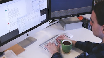 Bir kişi masa başında oturmuş dizüstü bilgisayar kullanıyor. Önünde bir fincan kahve ve elinde bir bilgisayar faresi vardır. Kişi her iki eliyle de klavyede yazı yazmaktadır. Yanındaki masada üzerinde delikler olan gümüş renkli dikdörtgen bir nesne var. Kişinin önünde içinde kahverengi bir sıvı olan yeşil bir kupa vardır. Dizüstü bilgisayar ekranında mavi bir arka plan var. Kişi klavyeye odaklanmıştır ve elleri yazma pozisyonundadır. Fare sağ elinin yanında konumlandırılmıştır.