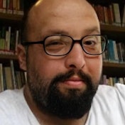 A mature man with a full beard and round glasses is looking directly at the camera. He is wearing a grey, long-sleeved shirt and a pair of dark-colored jeans. His hair is combed back away from his forehead, and a few strands rest on his forehead. His eyes are alert and his mouth slightly curved in a thoughtful expression. The man appears to be in his thirties, and his facial features are distinct. His beard is neatly trimmed and his glasses frame his face in a flattering way. He looks confident and ready to take on the world.