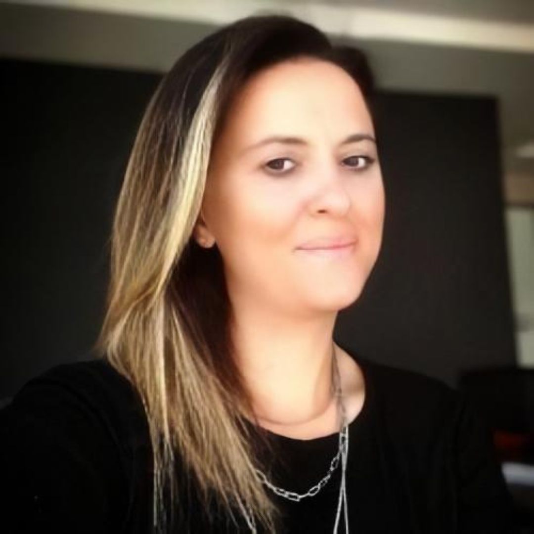 Uzun siyah saçları ve parıldayan gümüş kolyesiyle güzel ve genç bir kadın. Kıvrımlarını vurgulayan beyaz bir elbise giyiyor ve koyu renk gözleri uzun kirpiklerle çerçevelenmiş. Kendinden emin ve memnun görünüyor. Saçları ortadan ayrılmış ve omuzlarından aşağı dökülüyor ve kolyesinde tek bir parlak mücevher var. Neşe saçan sıcak ve davetkâr bir gülümsemesi var.