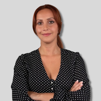 A young woman with bright red hair stands in front of a plain white background. She is wearing a black and white polka dot shirt with a V-neck. Her eyes are closed, and she has a slight smile on her face. Her arms are crossed in front of her, and her hands are slightly clasped together. She is wearing simple silver hoop earrings, and her hair is pulled back in a low ponytail. She looks relaxed and content, enjoying the moment.