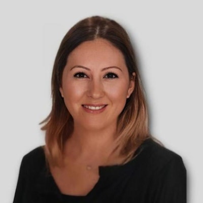 Genç bir kadın parlak fonlu bir stüdyoda duruyor ve kameraya gülümsüyor. Yüzünde dostça bir ifade var, dudakları hafifçe ayrılmış, gözleri parlıyor ve başı hafifçe yana eğilmiş. Uzun, siyah saçları geriye doğru toplanmış ve omuzlarından aşağı dökülüyor. Beyaz bir bluz, siyah bir etek ve siyah topuklu ayakkabı giyiyor. Elleri önünde kenetlenmiş, manikürlü güzel tırnaklarını gösteriyor. Rahat bir pozda duruyor, hoş ve kendinden emin bir hava yayıyor.