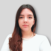 The woman in the image has long, dark hair that falls over her shoulders. She is wearing a white shirt with a collar and three-quarter length sleeves. Her expression is neutral. She is standing in front of a white wall. She is looking directly into the camera, her eyes slightly narrowed in concentration. Her lips are slightly parted and her eyebrows are raised slightly. Her hands are in front of her, her palms facing up. She looks confident and composed. Her posture is relaxed and her stance is confident. Her facial features are well-defined and her skin tone is a light tan.