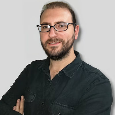 A middle-aged man wearing glasses and a full beard is facing forward with a serious expression on his face. He is wearing a gray, long-sleeve shirt with a black tie and is standing in a white-walled room with a light brown floor. His hair is dark brown and combed back, and his eyes are a brownish-green color. His glasses appear to be square-framed and have lenses that are slightly tinted. His facial features, such as the shape of his nose and his lips, are visible. He looks calm and composed, standing tall and proud.