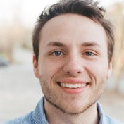 A young man is sitting in front of the camera with a warm smile on his face. He has a light complexion, with short, dark brown hair, a strong jawline, and light brown eyes. His attire is casual: a white t-shirt and jeans. He is looking directly at the camera, his gaze direct and inviting. His expression is one of contentment and joy, radiating outward to the viewer. His posture is relaxed but confident, as he sits with his hands on his lap. He appears to be in his late twenties, with a healthy and vibrant look to him. His presence is inviting and comforting, a perfect representation of the kind of joy that comes with a peaceful moment.