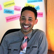 A man with a bright, inviting smile is looking directly at the camera. He has a light brown complexion, short, dark hair, and a neatly groomed beard. His eyes are twinkling with joy and his mouth is slightly open, showing off his perfect teeth. He is wearing a white collared shirt and black trousers, and appears relaxed and content. The background is blurry, adding to the focus on the man's face and smile. He looks happy and ready to take on the world.