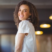 A cheerful woman with her head tilted slightly to the side and a warm smile on her face looks directly into the camera lens. Her short brown hair is neatly styled, and her beige eyes twinkle with delight. She is wearing a light blue and white striped shirt, and her lips are slightly parted in a joyful expression. Her hands are resting on her lap, and her body is relaxed in a comfortable pose. The bright sunlight highlights her features and the vibrant colors of her clothing. She looks relaxed and happy, her smile radiating joy and contentment.