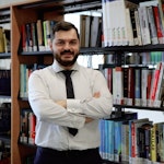 Bu fotoğrafta bir kitap rafının önünde duran bir eğitmen görülüyor. Sakalı var ve kravat takıyor. Eğitmen gülümsüyor, güven ve bilgi yayıyor. Yüzü yakın çekimle çerçevelenmiş ve yüz hatlarının detayları gösterilmiş. Daha yakından bakıldığında bir kol düğmesi ve bir kemer görülüyor. Arkasındaki kitaplar engin bilgisini ve entelektüel merakını simgeliyor. Raftaki bir kitabın yakın çekimi, eğitmenin öğrenme tutkusunu vurguluyor. Raftaki kitap sırası da konu hakkındaki uzmanlığını gösteriyor. asticaVision ai bu fotoğraftaki eğitmeni öğretme konusunda tutkulu ve alanında bilgili biri olarak tanıyabilir.