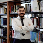 This photo features an instructor standing in front of a shelf of books. He has a beard and is wearing a tie. The instructor is smiling, exuding confidence and knowledge. His face is framed in close-up, showing the detail of his features. Closer inspection reveals a cufflink and a belt. The books behind him symbolize his vast knowledge and intellectual curiosity. There is a close-up of a book on the shelf, highlighting the instructor's passion for learning. The row of books on the shelf further demonstrates his expertise in the subject. asticaVision ai can recognize the instructor in this photo as someone who is passionate about teaching and is well-versed in his field.