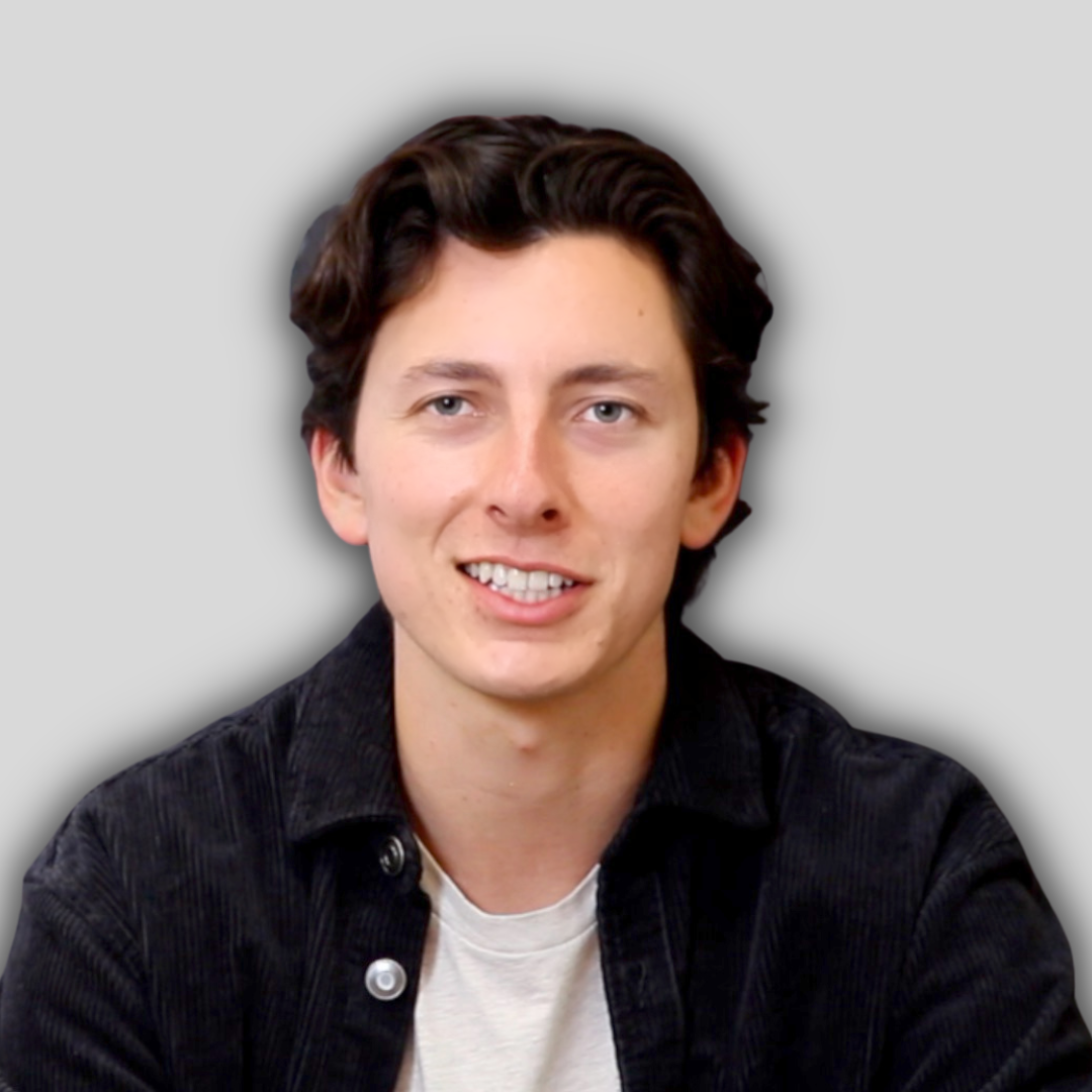 The man in the image is smiling with a wide, toothy grin. His eyes are bright and his cheeks are slightly rosy. He is wearing a white t-shirt, with his arms crossed casually. He is looking directly at the camera, his face relaxed and full of joy. His hair is cropped short and neat, and his skin is smooth and clear. He appears to be in his early twenties, and is in a bright, sunlit outdoor setting. The background is a vibrant, lush green, with the sun's rays peeking through the trees. The man's smile is contagious, and he exudes a sense of contentment and happiness.