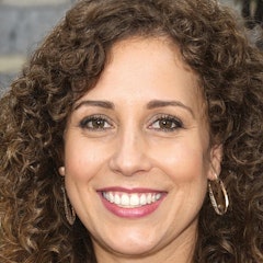 This close-up image shows a woman with a warm and inviting expression on her face. She has her eyes closed, as if she is savoring a moment of joy or contentment. Her lips are curved up into a wide, genuine smile, her teeth showing and her cheeks lifted. Her eyebrows are slightly raised, adding to her cheerful expression. Her long dark hair is swept away from her face, framing her features. Her skin is fair and her eyes are a deep brown. Her face has a soft, peaceful quality, radiating warmth and kindness.