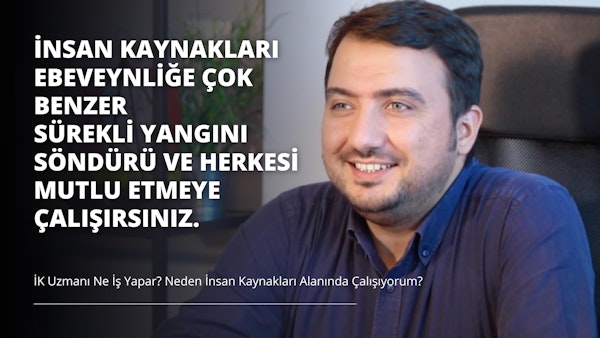 Sakallı bir adam kameraya gülümsüyor. Lacivert düğmeli bir gömlek giymiş ve arka planda yeşil bir bitki olan bir sandalyede oturuyor. Fotoğrafın sağ tarafında gri zemin üzerine beyaz bir harf ve siyah zemin üzerine üç beyaz harf var. Adam dostça bir ifadeyle doğrudan kameraya bakıyor. Yüzündeki saçlar düzgünce kesilmiş ve bir gözlük takıyor. Arka plandaki ışık yüzünü vurguluyor ve çerçevedeki renkleri aydınlatıyor. Fotoğraf bir neşe ve mutluluk anını yakalıyor.