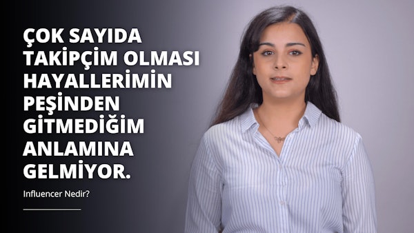 Uzun siyah saçlı bir kadın, mavi çizgili beyaz bir gömlek giymiş olarak beyaz bir arka planın önünde durmaktadır. Kolyesi takılı ve gömleğinin önünde küçük beyaz bir 'O' harfi görülüyor. Saçları yana doğru ayrılmış ve omuzlarına kadar iniyor. Kolye, küçük bir kolye ucu olan altın bir zincir ve resmin sağ tarafında kolyenin yakın çekimi var. Rahat bir duruşu var ve doğrudan kameraya bakıyor.