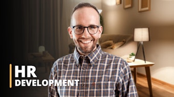 A man wearing glasses and a checkered shirt is in the foreground explaining a human resources development course. There is a house in the background. Gray wall and coffee table are visible. HR DEVELOPMENT is written above the image.