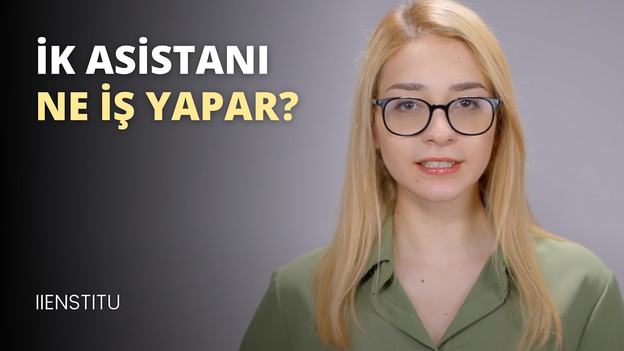 Gözlüklü ve yeşil gömlekli bir kadının yakın plan portresi. Ciddi bir ifadeyle dosdoğru karşıya bakıyor. Gözlüklerinin arkasından gözleri görünüyor ve ağzının köşesinde küçük bir beni var. Saçları düzgün bir topuzla arkadan bağlanmış ve yüzü gözlük ve gömleği tarafından çerçevelenmiş. Gömleği yeşil renkte ve üzerinde küçük beyaz puantiyeler var. Basit bir kolye ucu olan ince gümüş bir kolye takıyor. Gömleğinin altından göğsü görünüyor ve kolları iki yanında. Arka planda beyaz harflerle yazılmış siyah beyaz bir tabela ve siyah zemin üzerine beyaz bir harf var.