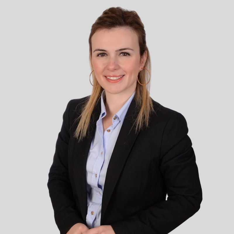 Bu, profesyonel iş kıyafeti giyen genç bir kadının fotoğrafı. Mavi duvarlı ve kahverengi halılı iyi aydınlatılmış bir odada duruyor. Kadının elleri cebinde ve duruşu kendinden emin ve dengeli. Koyu renk saçları düzgün bir şekilde toplanmış ve yüzü hafifçe makyajlanmış. Lacivert bir takım elbise ceketi, altında beyaz bir bluz ve lacivert bir pantolon giyiyor. Siyah ayakkabıları cilalı ve ona sofistike bir görünüm veriyor. Gururlu ve kendinden emin görünüyor ve ifadesi sakin ve odaklanmış.