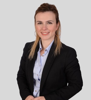 This is a photo of a young woman wearing a professional business suit. She is standing in a well-lit room with a blue wall and brown carpet. The woman has her hands in her pockets, and her posture is confident and poised. Her dark hair is styled in a neat updo, and her face is lightly made-up. She is wearing a navy blue suit jacket with a white blouse underneath, and navy blue trousers. Her black shoes are polished and give her a look of sophistication. She looks proud and self-assured, and her expression is calm and focused.