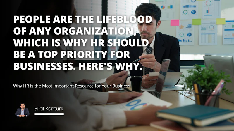 A man in a suit stands in front of a woman and talks to her. He is wearing glasses and a smart suit and holds a pen in his hand. A black plastic cup with a lid sits nearby. A plant in a pot is on a desk in the background. A blurry image of a stack of books also sits in the background. A man with glasses and a beard is visible to the side. A group of people are sitting at a table further in the background. In the foreground is a white letter 'O' on a black background. A close-up of a hand is seen holding a white plate.