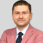 Özel dikim siyah takım elbise giyen ve kravat takan yakışıklı, iyi giyimli bir adam uzun boylu ve kendinden emin bir şekilde duruyor. Takım elbisesinin altında açık renkli düğmeli bir gömlek vardır ve güçlü çene hattı özenle bakımlı yüz saçlarıyla vurgulanmıştır. Duruşu rahattır ve elleri cebinde durur, bu da ona kendine güvenen bir hava verir. Koyu renk saçları taranarak şekillendirilmiş ve gözleri kararlı bir bakışla doğrudan ileriye bakıyor.