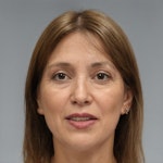 Bu, açık kahverengi saçlı bir kadının yakın çekim fotoğrafı. Gözleri kapalı ve yüzünde hafif bir gülümseme var. Yüz hatları yumuşak bir şekilde aydınlatılmış, bu da ona sıcak ve huzurlu bir görünüm veriyor. Yakasında ve kollarında ince siyah bir kenarlık olan beyaz bir üst giyiyor. Dudakları açık pembe bir renge boyanmış ve koyu kahverengi bir kolye takıyor. Cildi kusursuz ve kaşları özenle alınmış. Arka plan bulanık ve görüntünün sıcaklığına katkıda bulunan altın rengi bir tona sahip. Kadın bir huzur ve dinginlik havası yayıyor ve güzelliği şüphe götürmez.