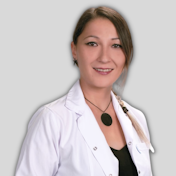 A young woman is standing in a white coat, looking away from the camera. She has long, dark brown hair and her eyes are drawn in a serious, focused expression. Her white coat is buttoned up to her neck and she is wearing a pair of black trousers. Her hands are clasped in front of her, and her posture is upright and confident. She has an air of sophistication and calmness. She looks confident and poised, with a sense of poise and determination. She looks ready to take on any challenge.