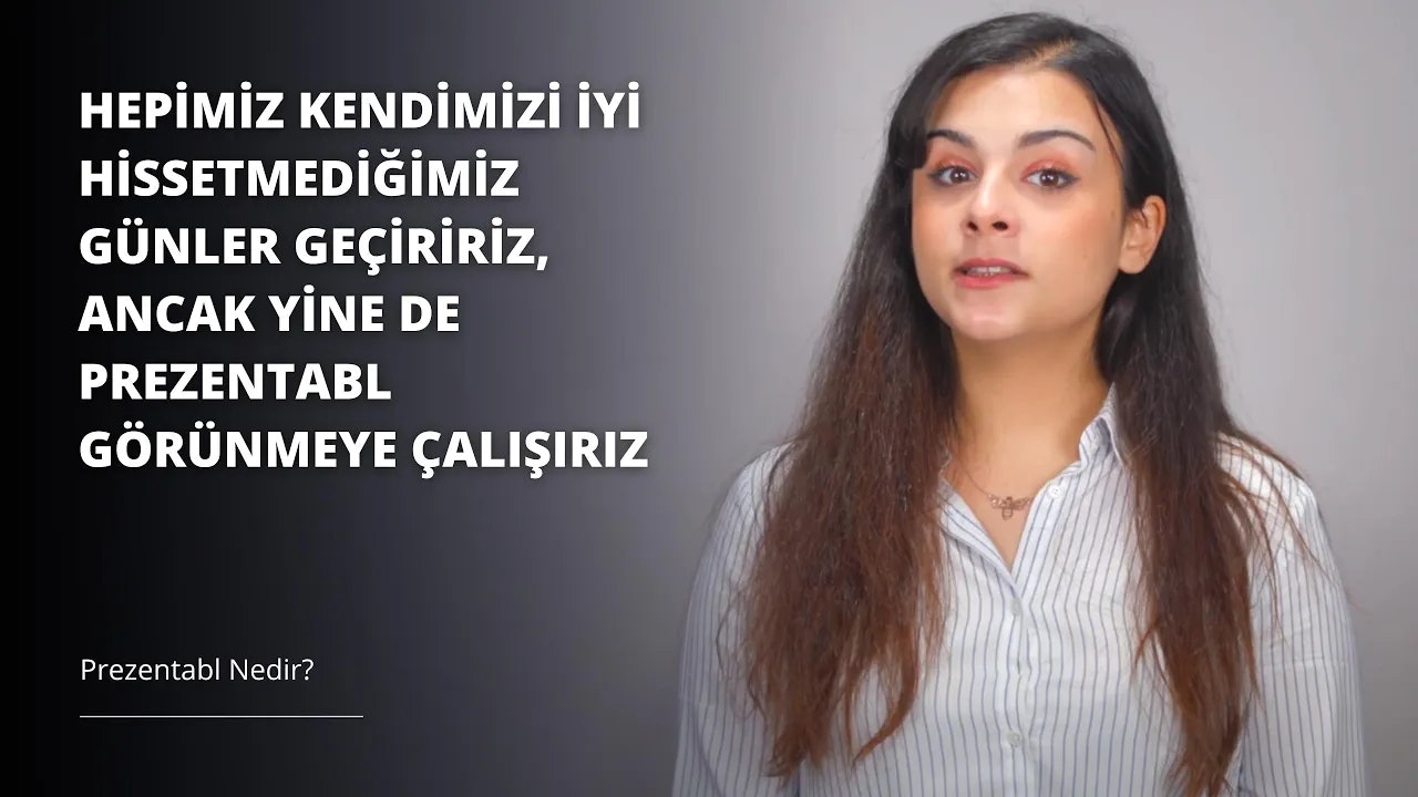 Resimde uzun, koyu kahverengi saçlı genç bir kadın görülüyor. Sıcak, hoş bir ifadesi var ve bakışları hafifçe yukarıya ve sola doğru yönelmiş. Saçları hacimli ve omuzlarının etrafında hafifçe hareket ederek yüzünü çerçeveliyor. Cildi pürüzsüz ve hafif bronzlaşmış, burnunda ve yanaklarında birkaç çil var. Kıyafetleri sade ve minimalist; soluk pembe bir gömlek ve koyu mavi bir kot pantolondan oluşuyor. Arka planda bir miktar sarı ve turuncu olan ışık dolu bir odada duruyor. Genel izlenim sıcaklık, huzur ve memnuniyetten oluşuyor.